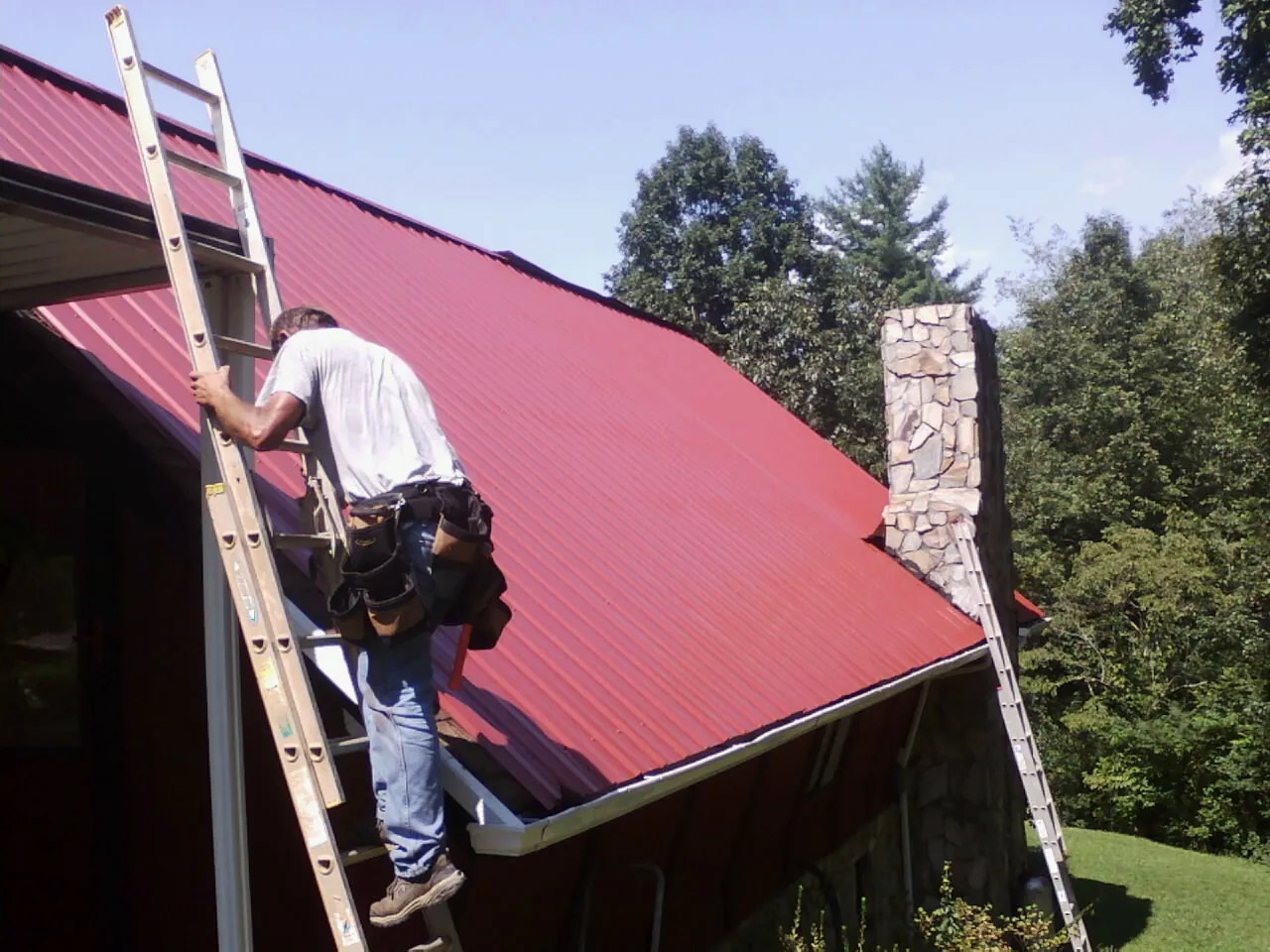 Roofer on a ladder beside a red metal roof and stone chimney — Soffit & Fascia Repair by Roofing Brothers in Saint Clairsville
